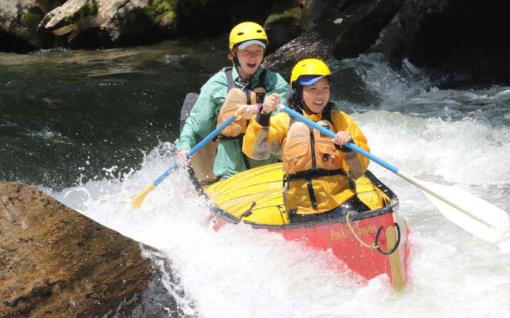 two students wearing safety gear paddle a canoe navigate whitewater in north carolina on an outward bound trip